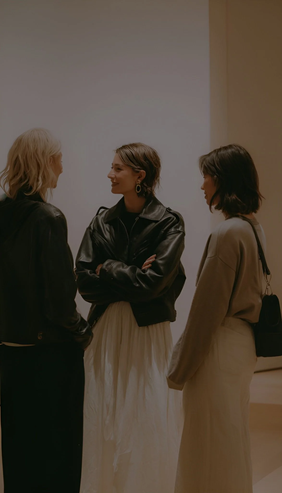 Three women having a conversation indoors against a blank wall.