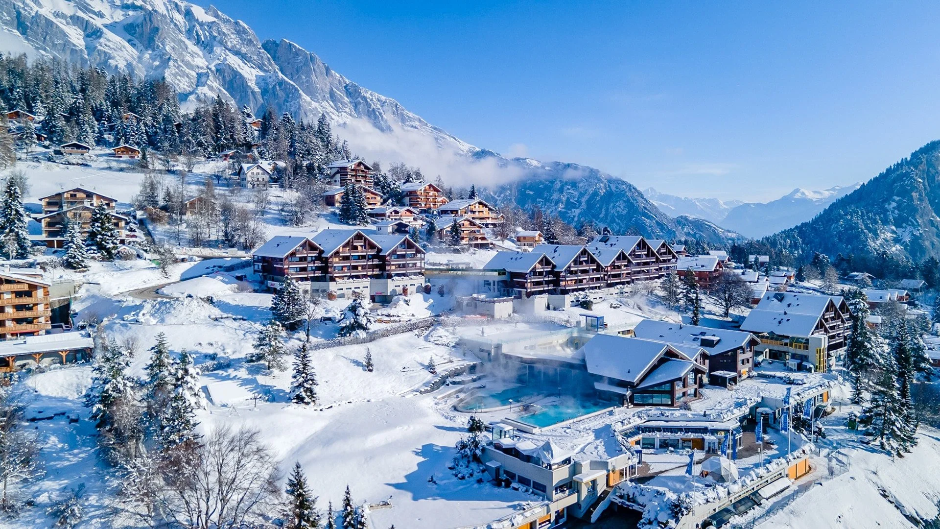 Village alpin enneigé avec des chalets en bois, montagnes en arrière-plan, et un spa avec piscine dans la neige, sous un ciel bleu clair.