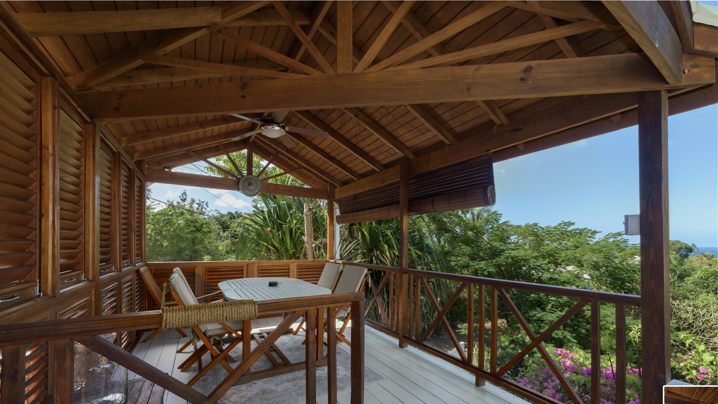 Wooden balcony with a table and chairs, ceiling fan, surrounded by greenery and trees, bamboo blinds, partial view of the landscape and ocean in the distance.
