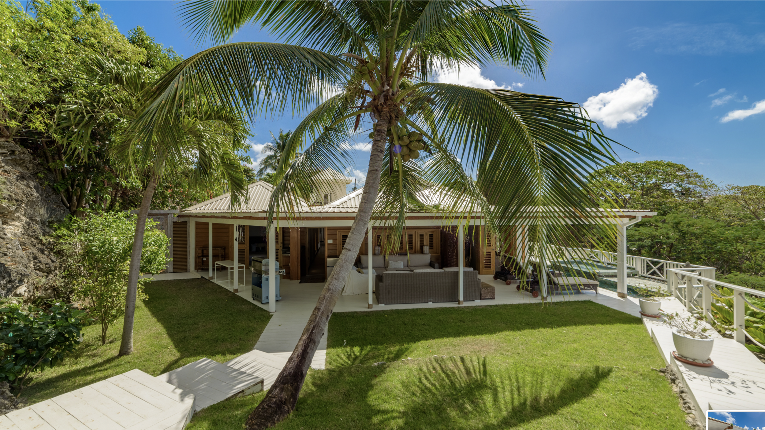 A tropical house with a white roof and wooden accents, situated in a lush green garden with palm trees. The house has an outdoor patio with seating and a grill, overlooking a backyard and surrounded by tropical forest.