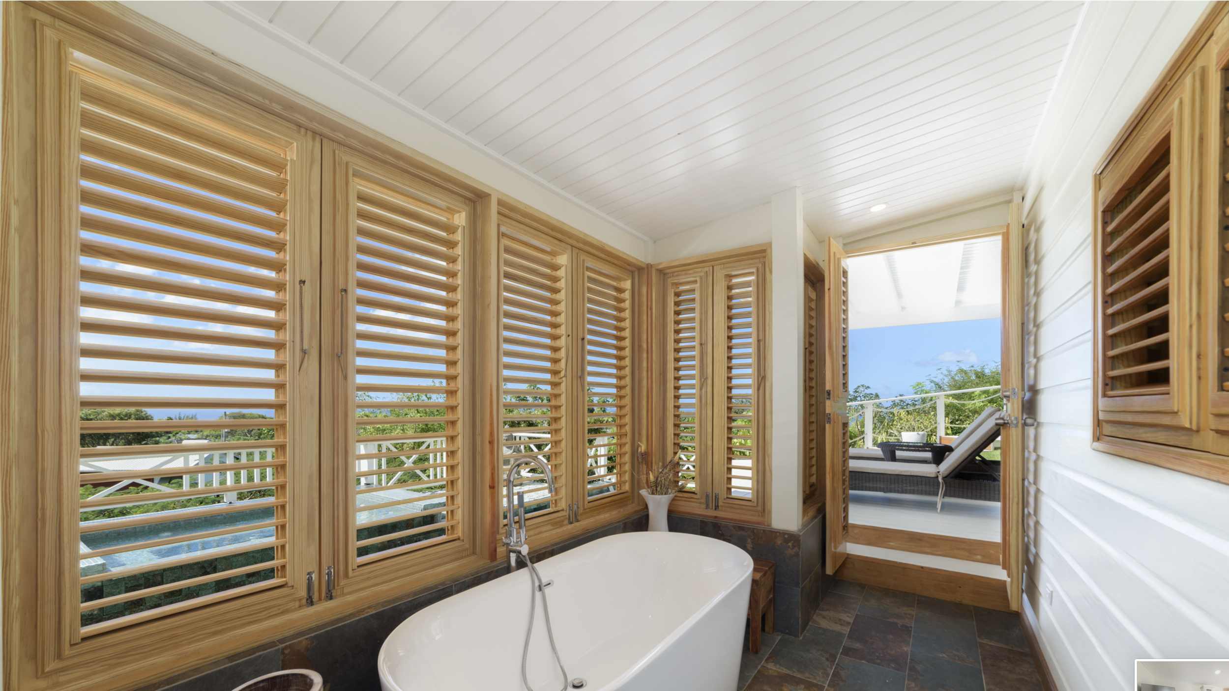 Bathroom with wooden shutters on windows, a white bathtub, a small side table with a vase, and a door leading to an outdoor patio with lounge chairs and a view of greenery and blue sky.