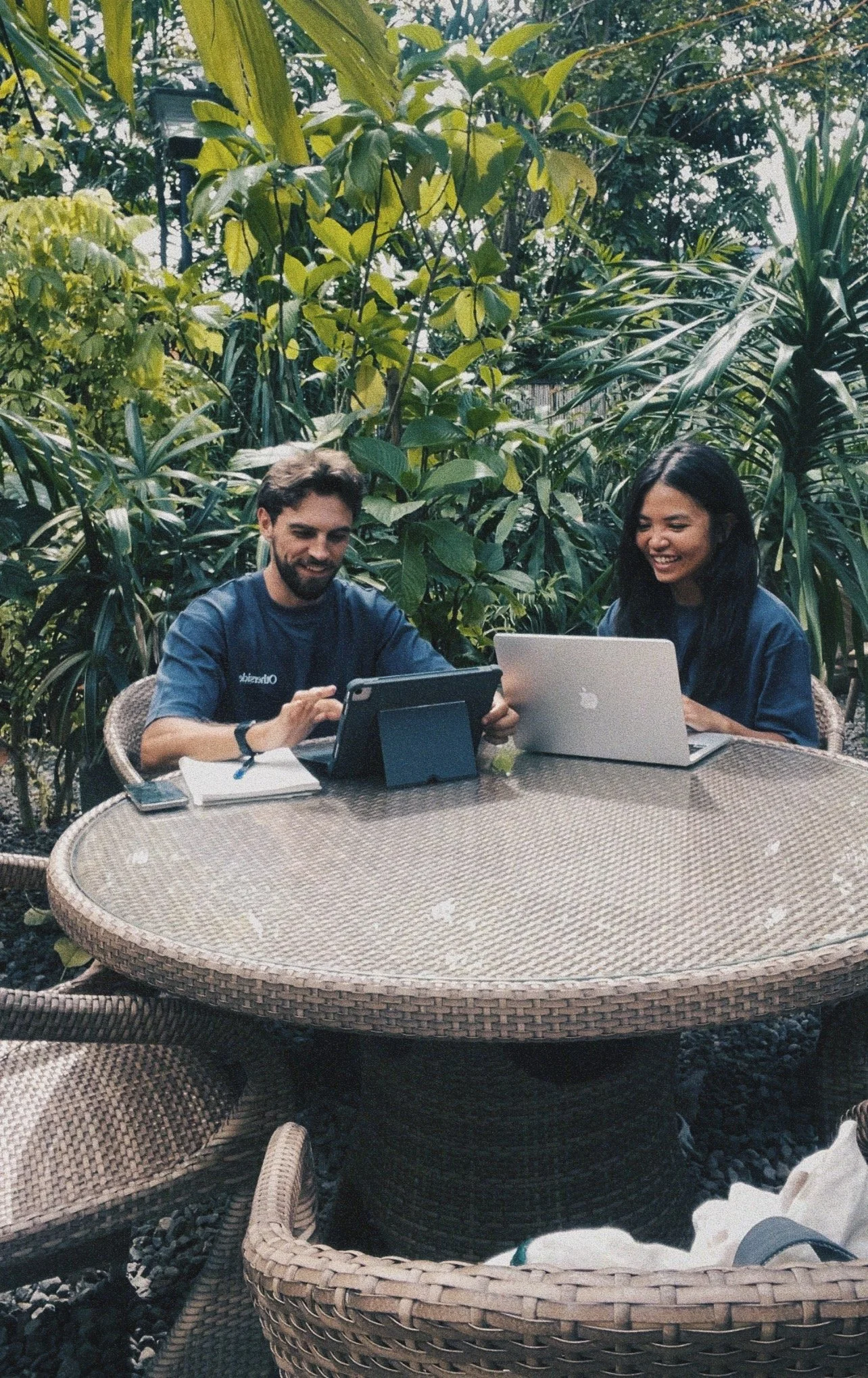 Two people sitting at a round wicker table outdoors, working on laptops, surrounded by dense green foliage.
