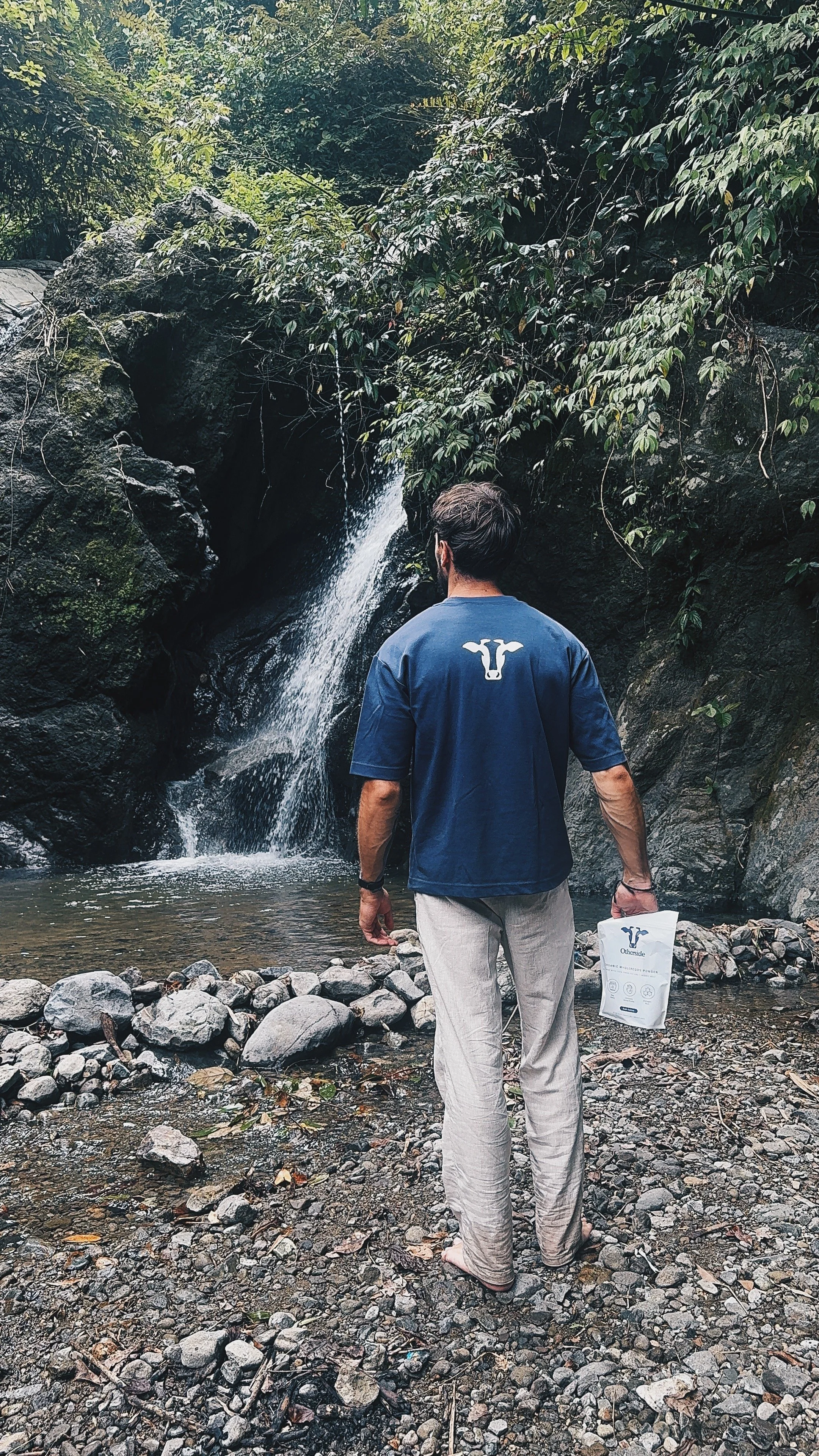 A man with dark hair and a beard standing on a rocky riverbank, facing a small waterfall surrounded by lush green foliage, holding a white paper bag with a company logo.