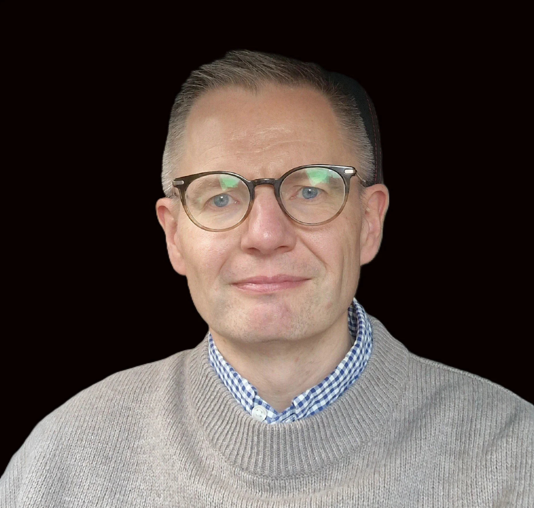Close-up of a middle-aged man with light blue eyes, gray hair, wearing glasses, a blue checkered shirt, and a beige sweater, smiling slightly against a plain black background.