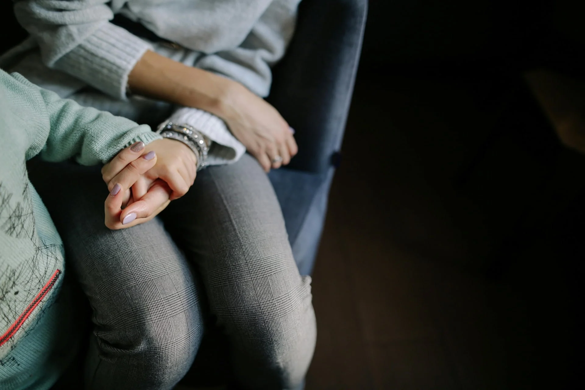 Two people holding hands, one wearing a light green sweater and the other wearing a grey sweater, sitting on a dark surface in a dark room.
