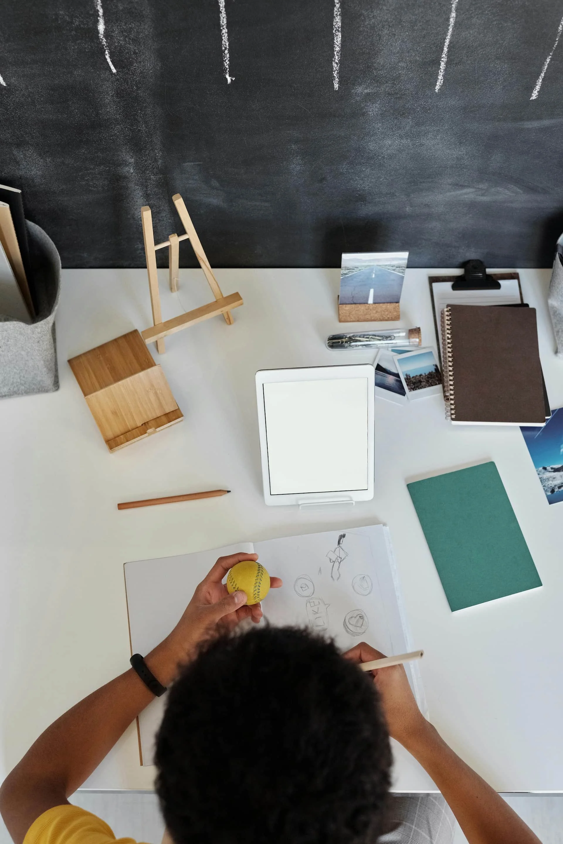 Top-down view of a person sketching with a pencil on a notebook at a cluttered desk, holding a yellow tennis ball. The desk has various items including a tablet, notebooks, photographs, a calendar, a pen, a small easel, and a blackboard or wall behind the desk.