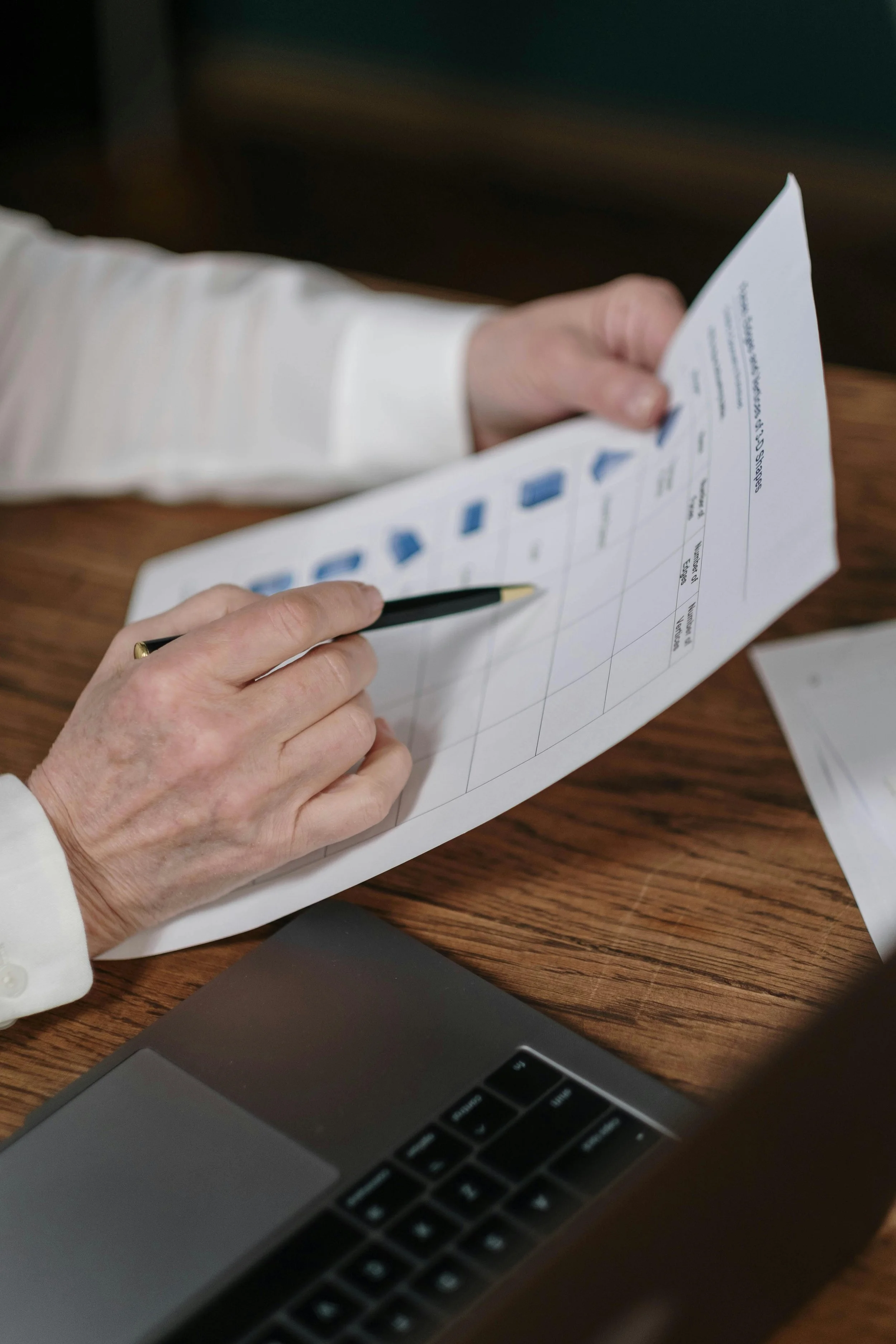 A person holding a document and pointing with a pen over a table with a laptop.