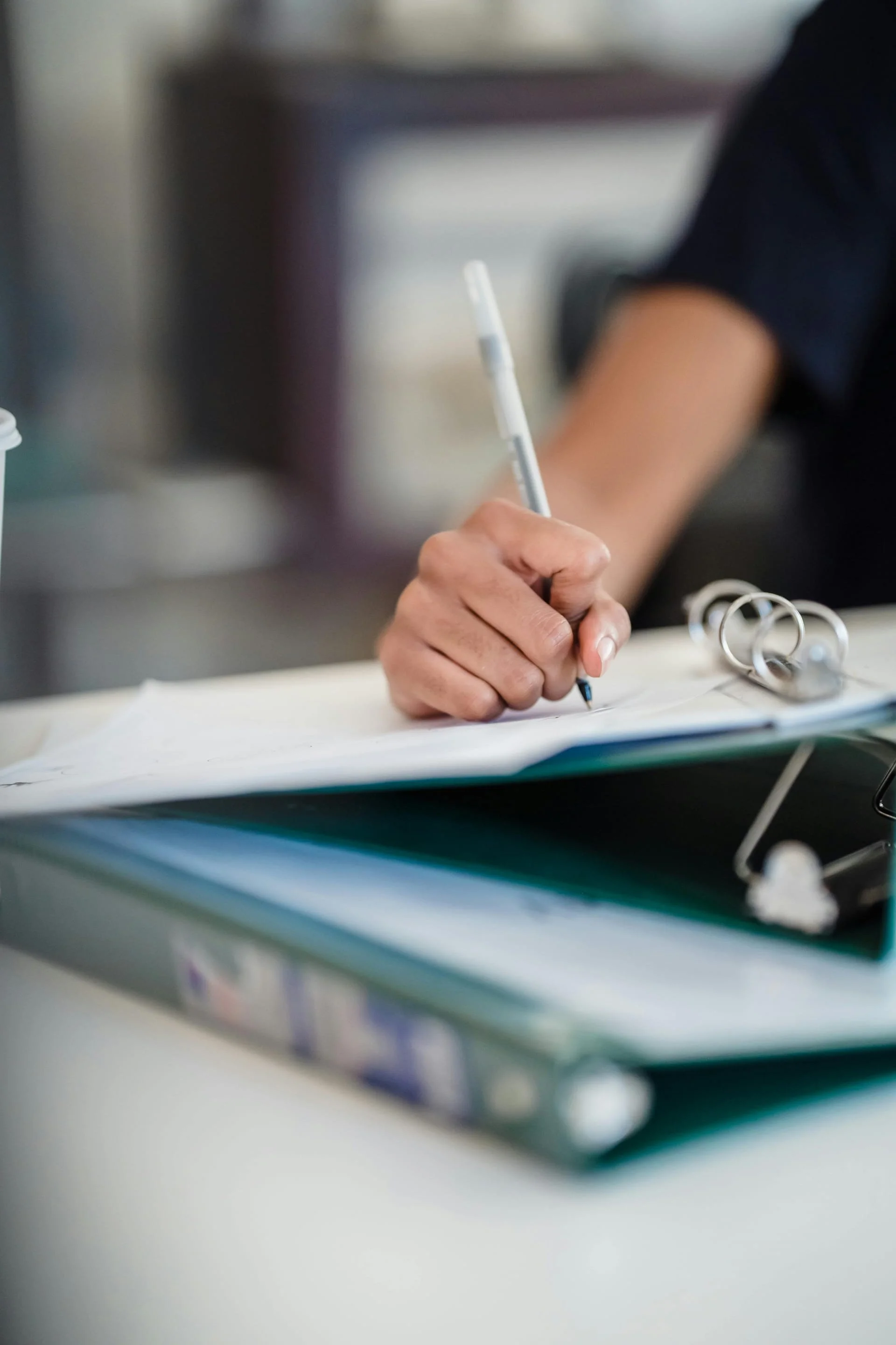 Person writing on a notepad with a pen, surrounded by a binder and documents on a desk.
