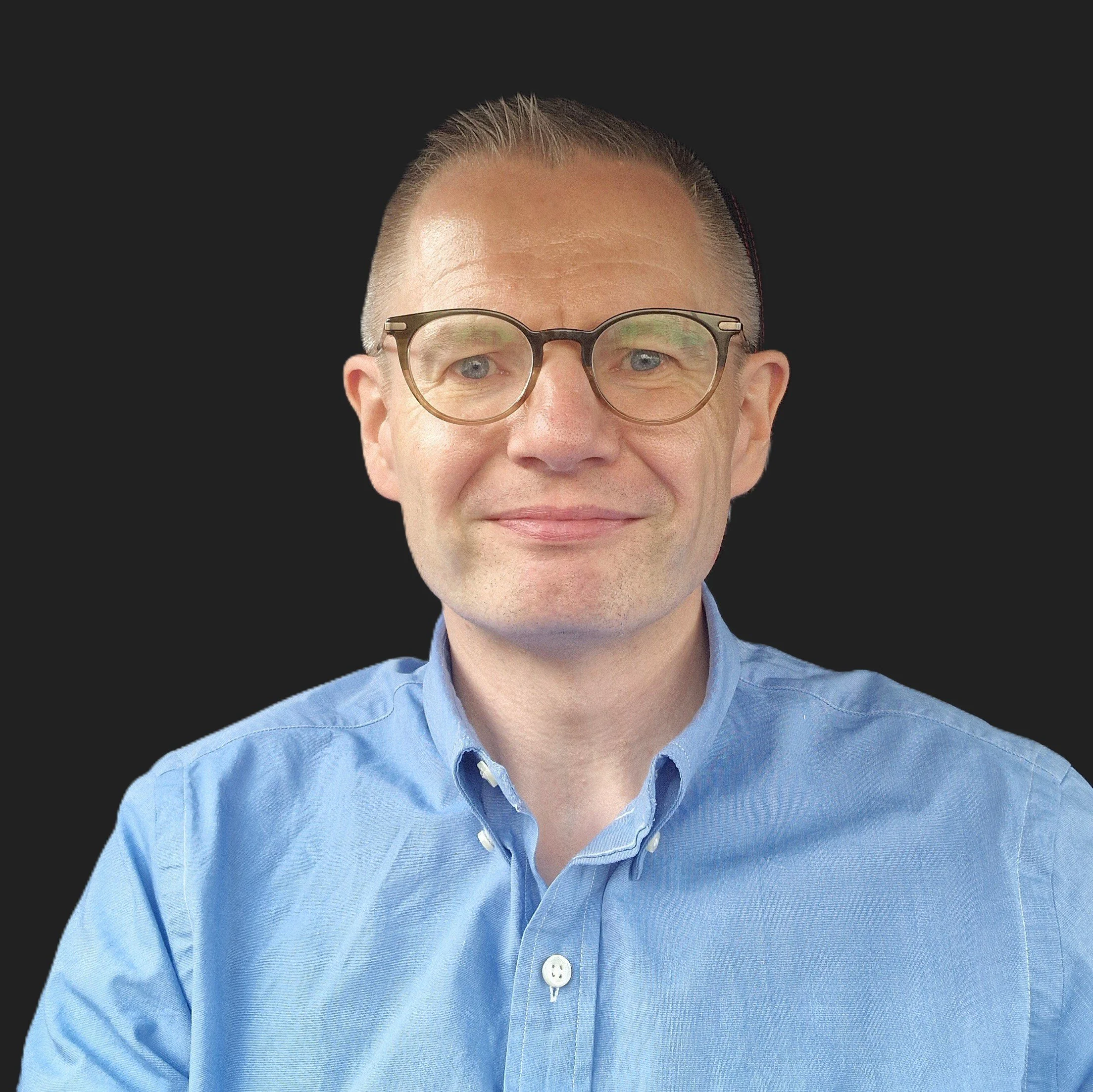 Close-up of a middle-aged man wearing glasses and a light blue collared shirt, with a black background.