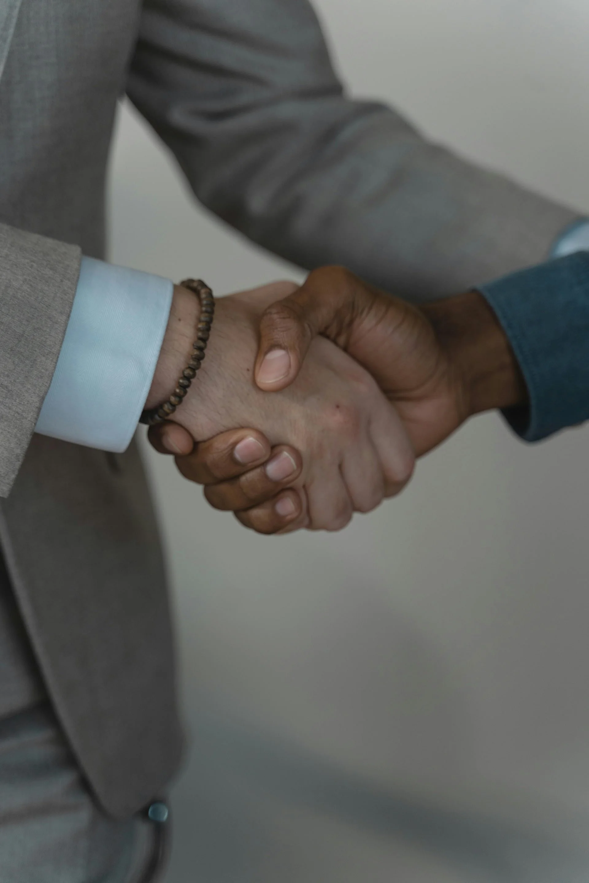 Close-up of two people shaking hands, one person wearing a gray suit and a light blue shirt, and the other person wearing a dark blue long-sleeve shirt. The background is plain and out of focus.