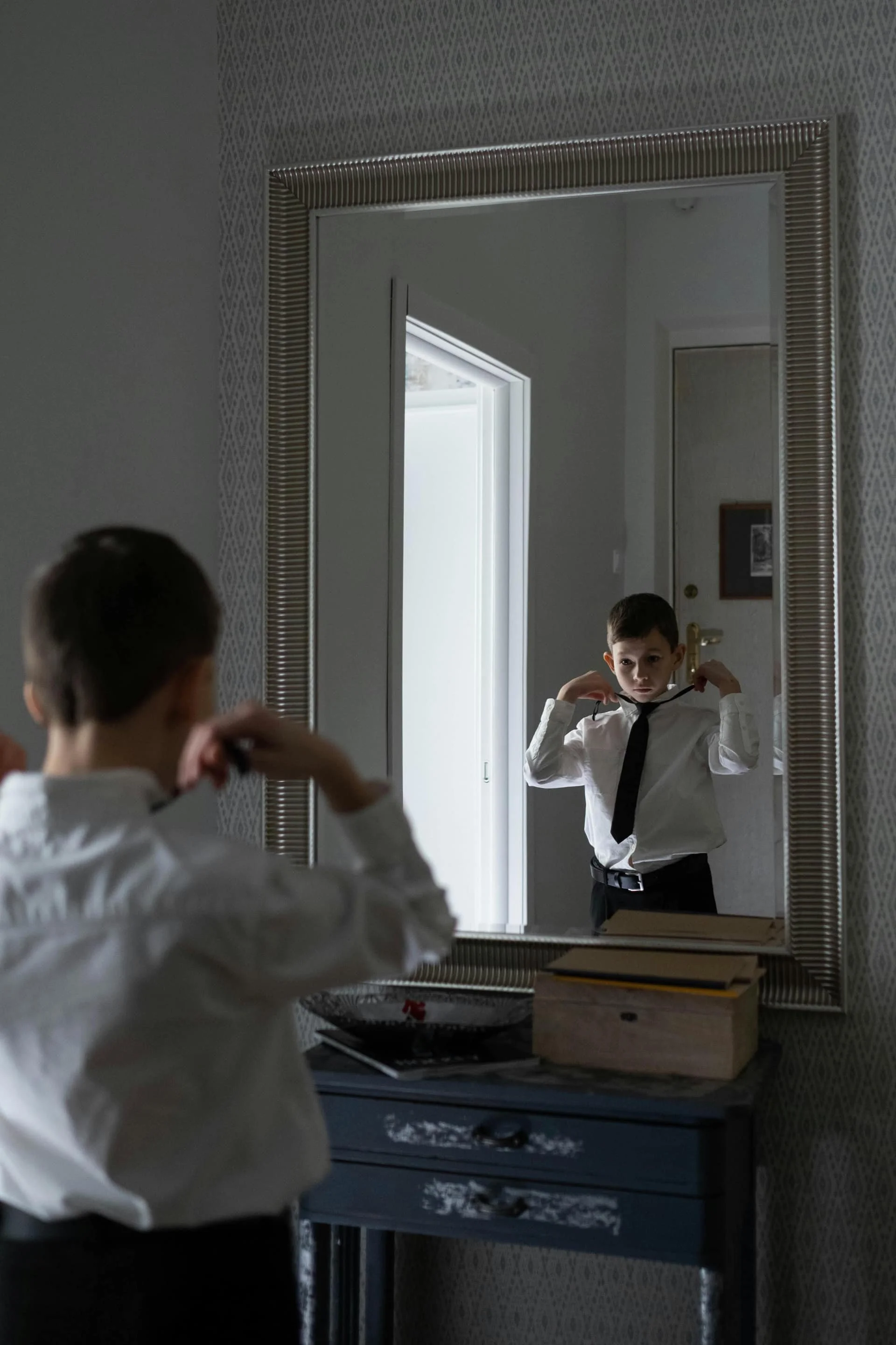 A young boy in a white shirt and black tie looks at himself in a mirror while adjusting his tie. The mirror's frame is decorative, and there is a blue dresser with some books and a bowl on top in front of the mirror. Light enters through an open door behind the boy.