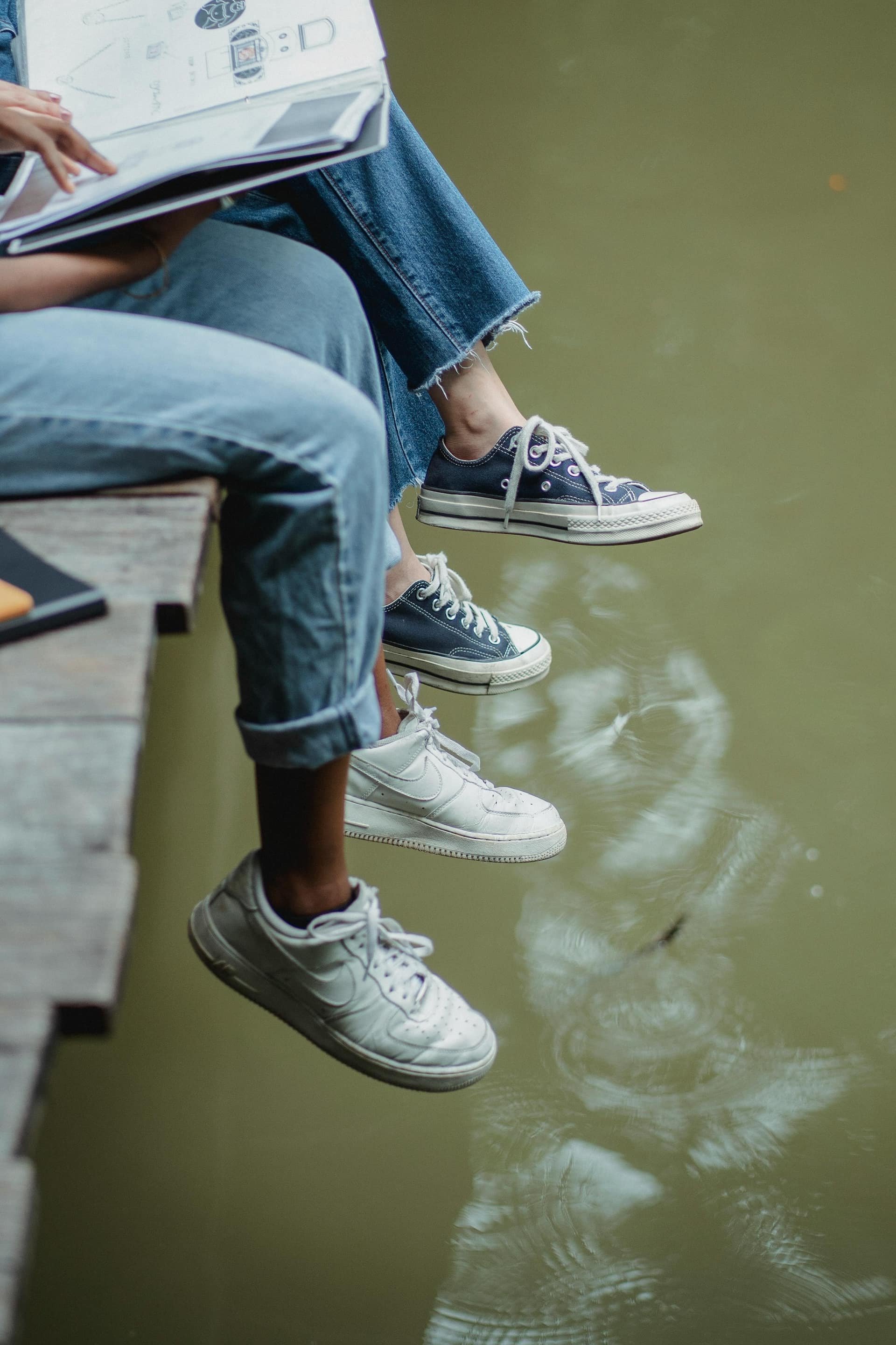 Three people sitting on a dock with their feet dangling over the water, wearing sneakers.