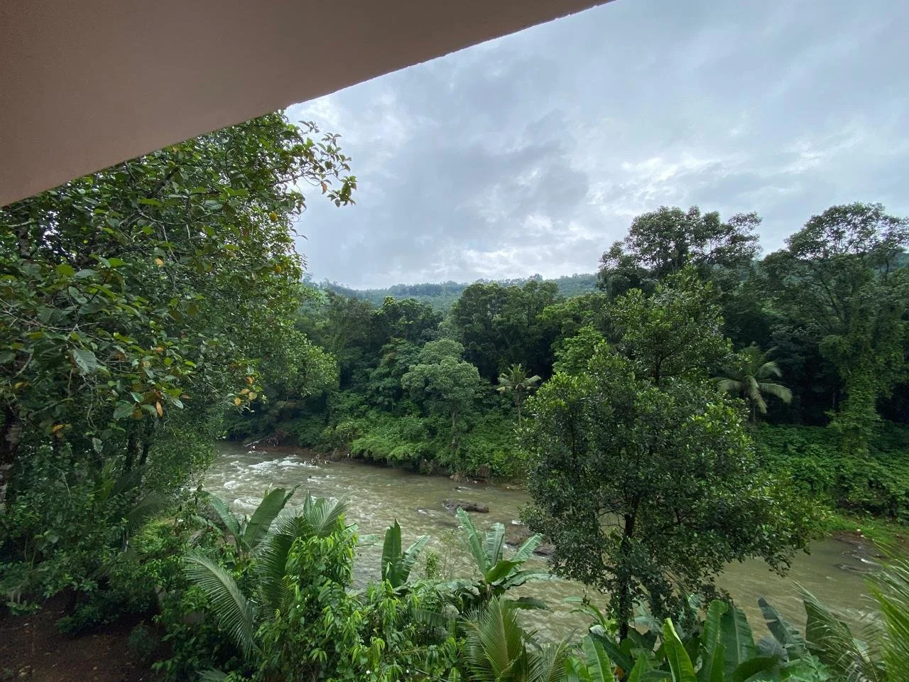 View of a river flowing through lush green tropical forest under a cloudy sky, seen from under a structure's overhang.
