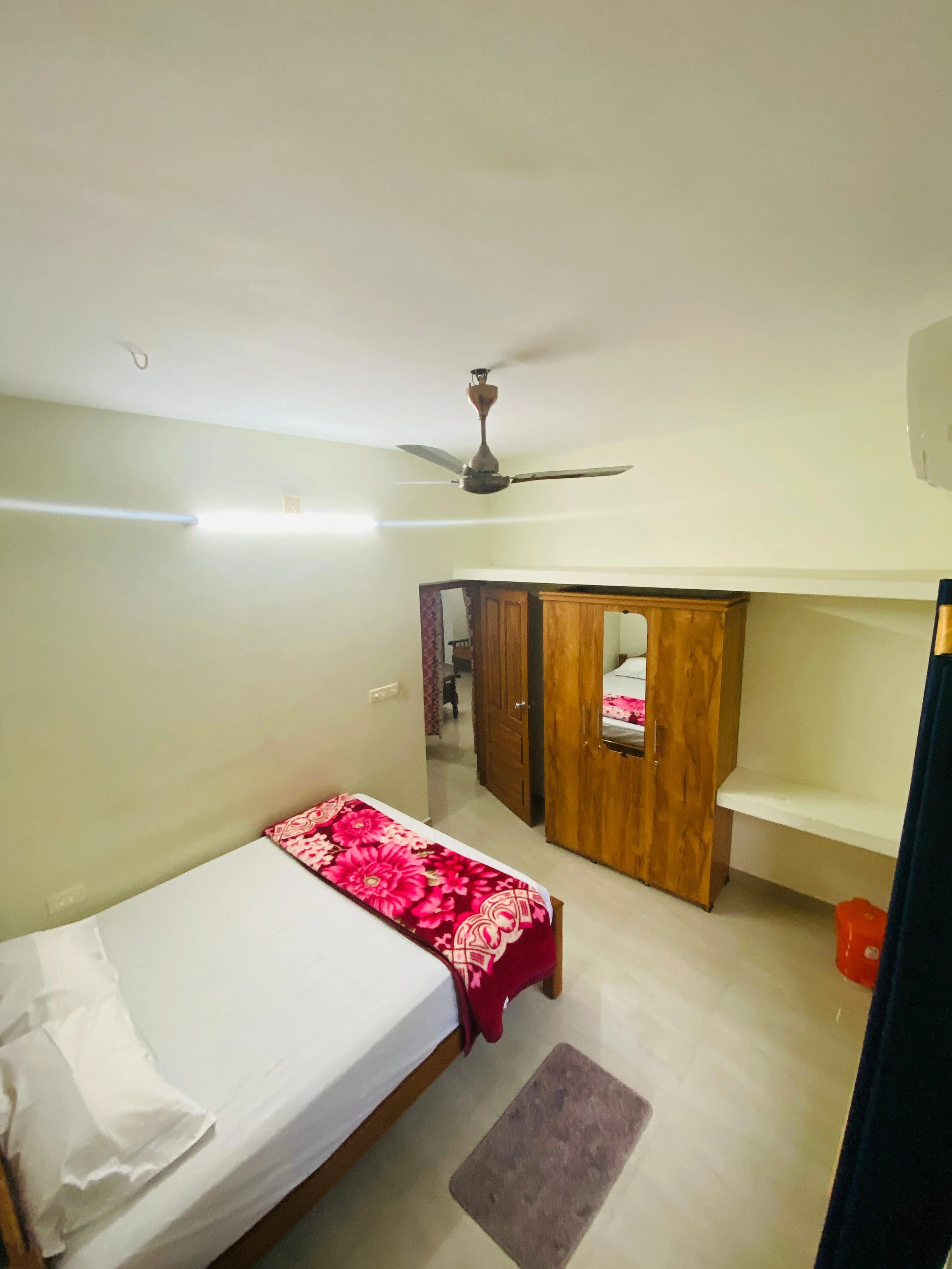 A simple bedroom with a bed, wooden wardrobe, wall mirror, ceiling fan, small rug, and minimal decor.