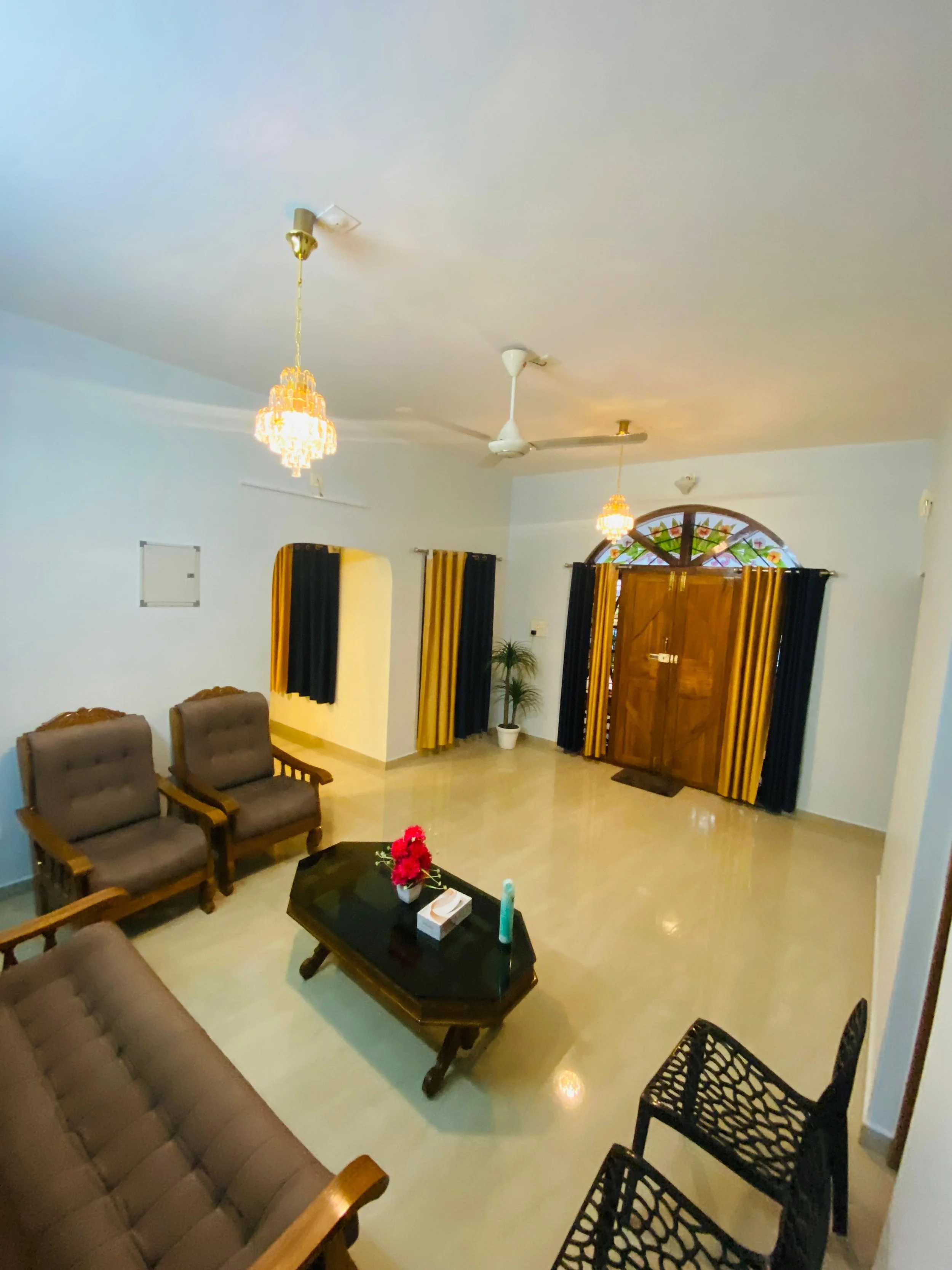 Living room with wooden front door, black and yellow curtains, ceiling fans, chandelier lights, brown upholstered chairs, a black coffee table with flowers and tissue box, and a small potted plant.