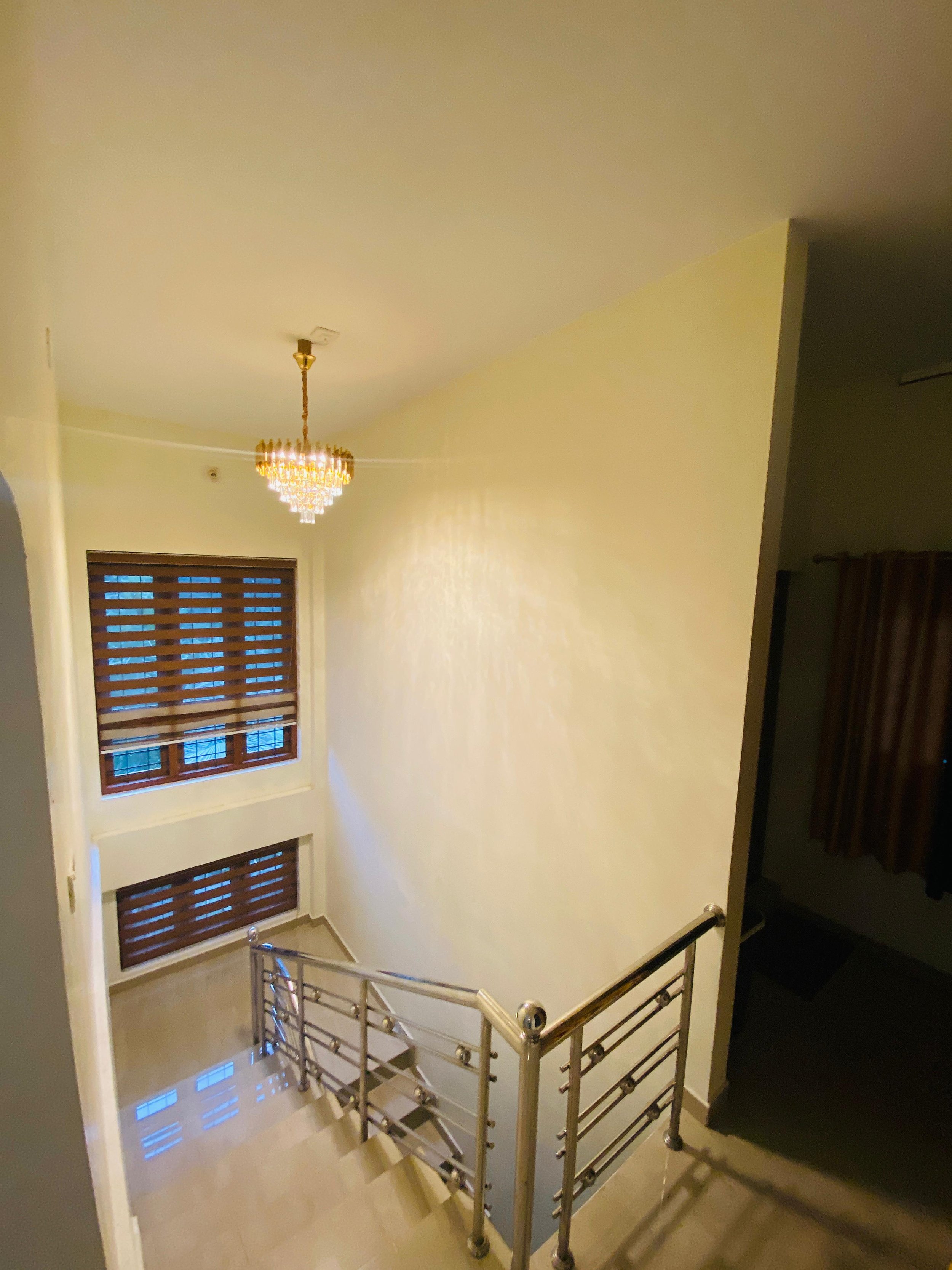Interior view of a staircase area with a chandelier, wooden blinds on windows, marble flooring, and a balcony railing.