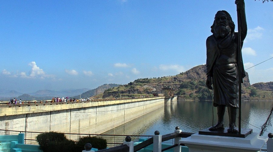 A large statue of a man with a beard standing near a dam, holding a staff, with a body of water and hills in the background.