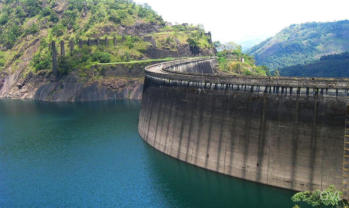 A large dam with a curved concrete wall across a body of water, with green hills and mountains in the background.