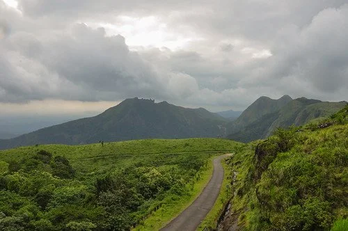 A winding road running through green lush hills with mountains in the background under a cloudy sky.