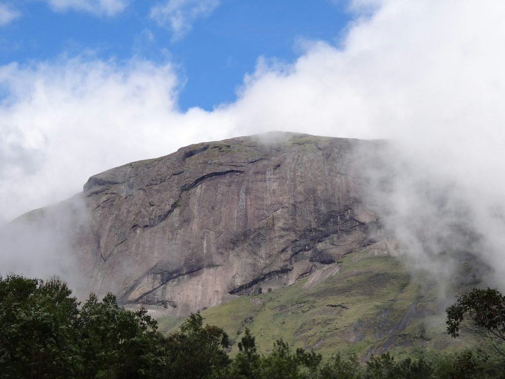 A large mountain with cloud cover, green vegetation at its base, and a bright blue sky with scattered white clouds.