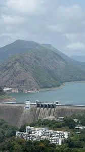 A mountain range next to a body of water with a dam and some buildings in the foreground.