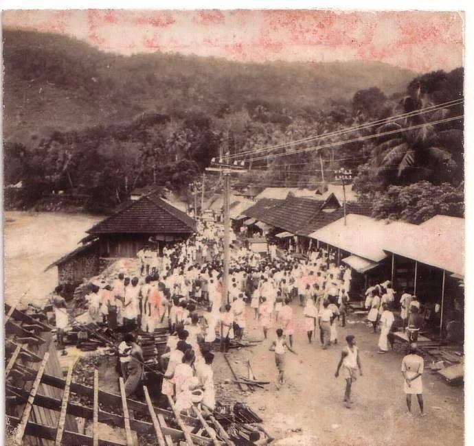 Historical black and white photo of a bustling train station or marketplace in a rural area with many people, wooden buildings, train tracks, and hilly landscape in the background.