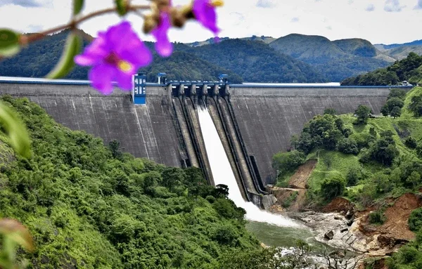 A large dam with water spilling over its gates, surrounded by green hills and mountains in the background.