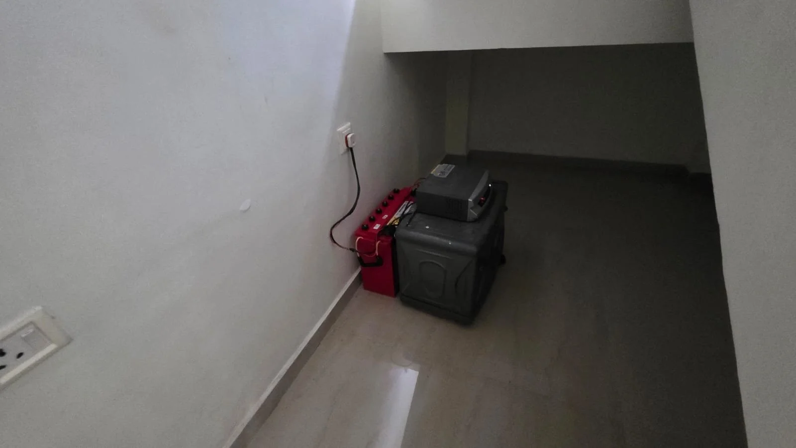 A corner of a room with a white wall, household electrical outlets, and a small rack with electronic devices including a black receiver and a red battery pack connected by cords.