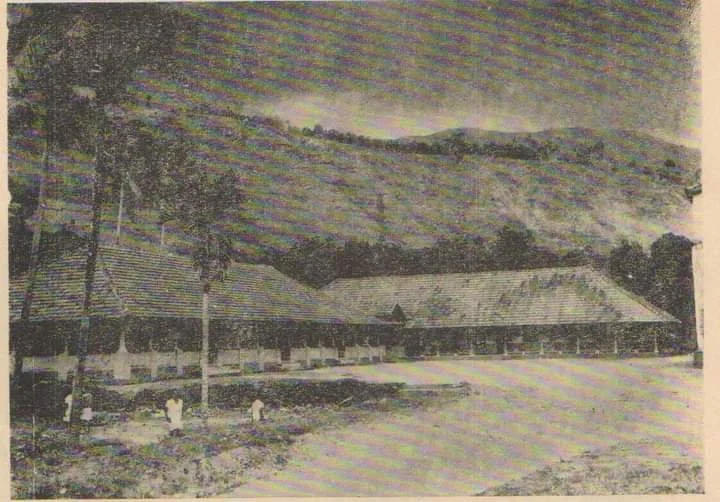 Historical black and white photograph of a long building with a sloped roof, situated in a rural area with a dirt road in front, with mountains in the background.