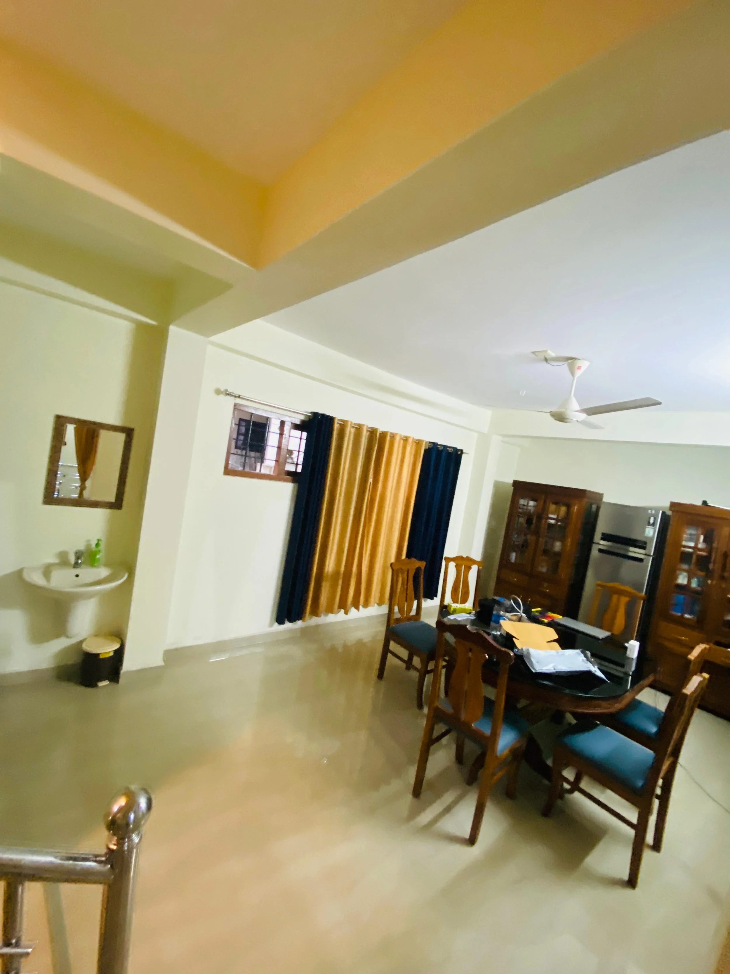 A living room with a dining table and chairs, wooden cabinets, curtains, a ceiling fan, and a small sink with a mirror on the wall.