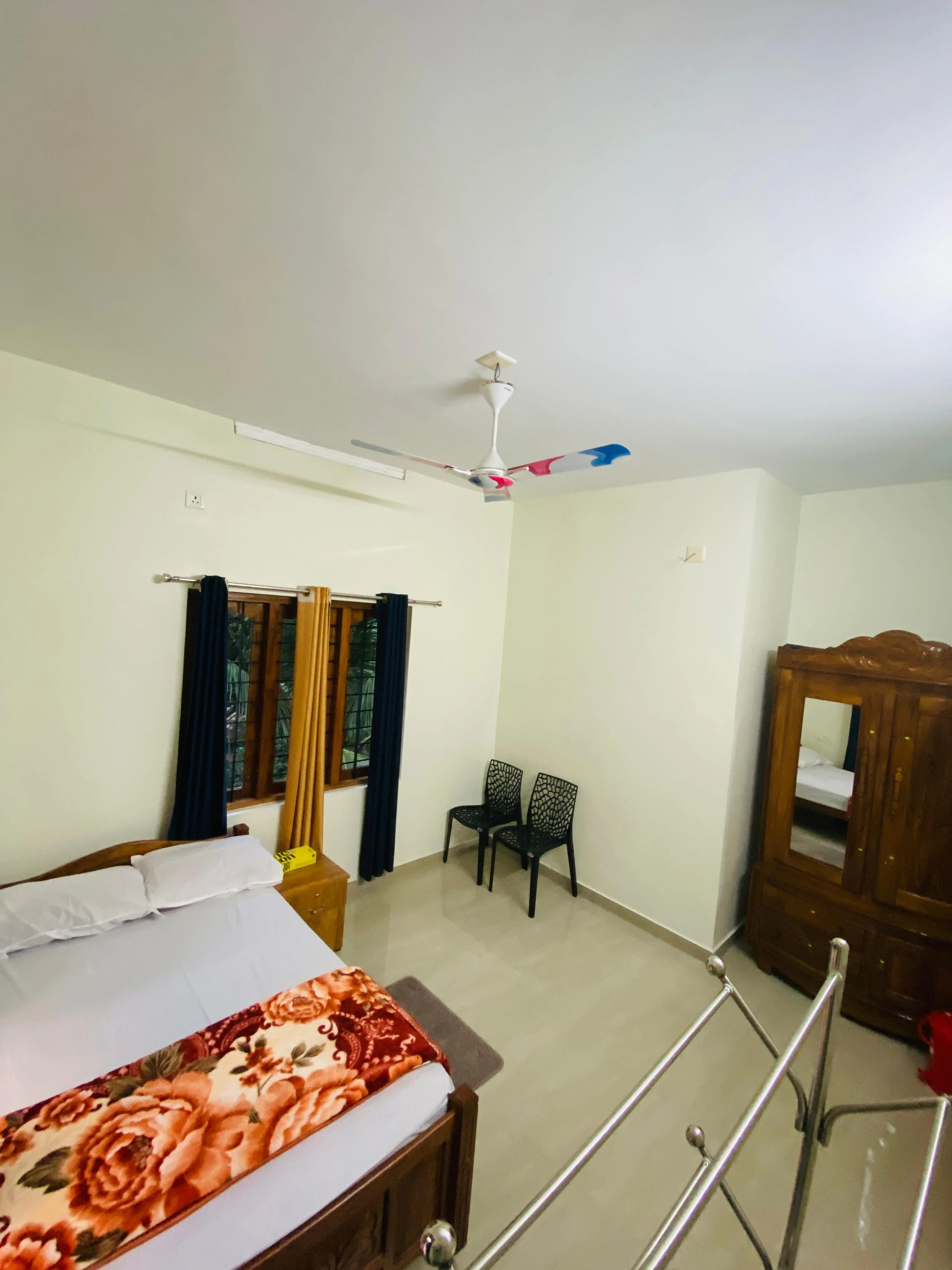 A bedroom with cream-colored walls and tiled floor, featuring a bed with a floral blanket, a wooden nightstand, two black patterned chairs, a large wooden wardrobe with mirror, a window with bars and curtains, and a ceiling fan with red, white, and b