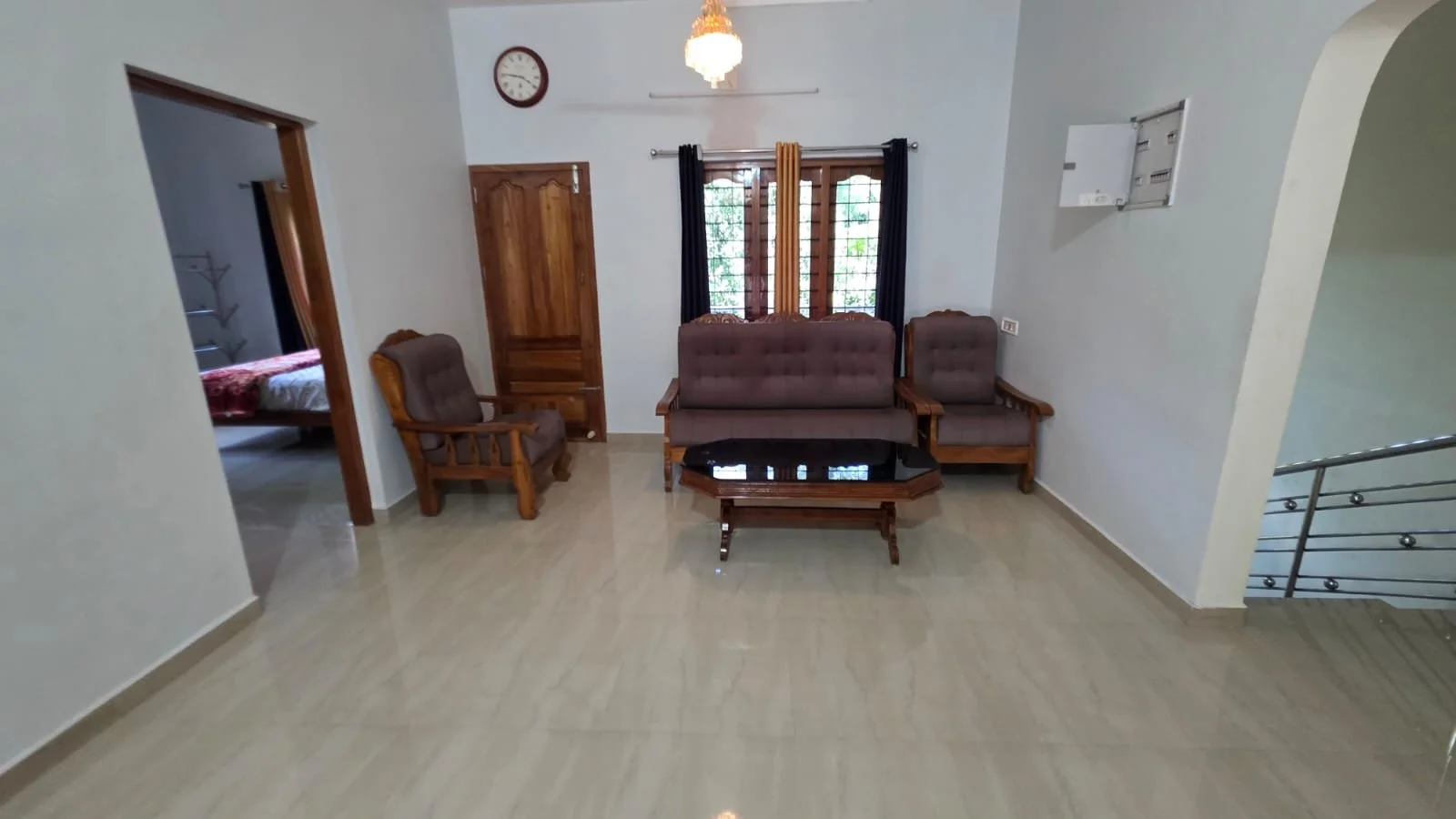 Living room with brown wooden furniture, including a sofa, two armchairs, and a coffee table with a black glass top. There are windows with black curtains, a wall clock, and a chandelier lighting fixture.