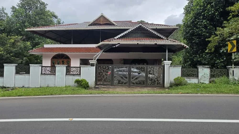 A house with a white fence and a decorative metal gate, surrounded by greenery and trees, situated next to a paved road.