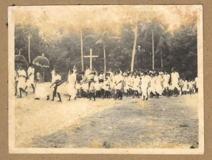 Children and adults participating in a religious or community event outdoors, standing in front of a cross with palm trees in the background.