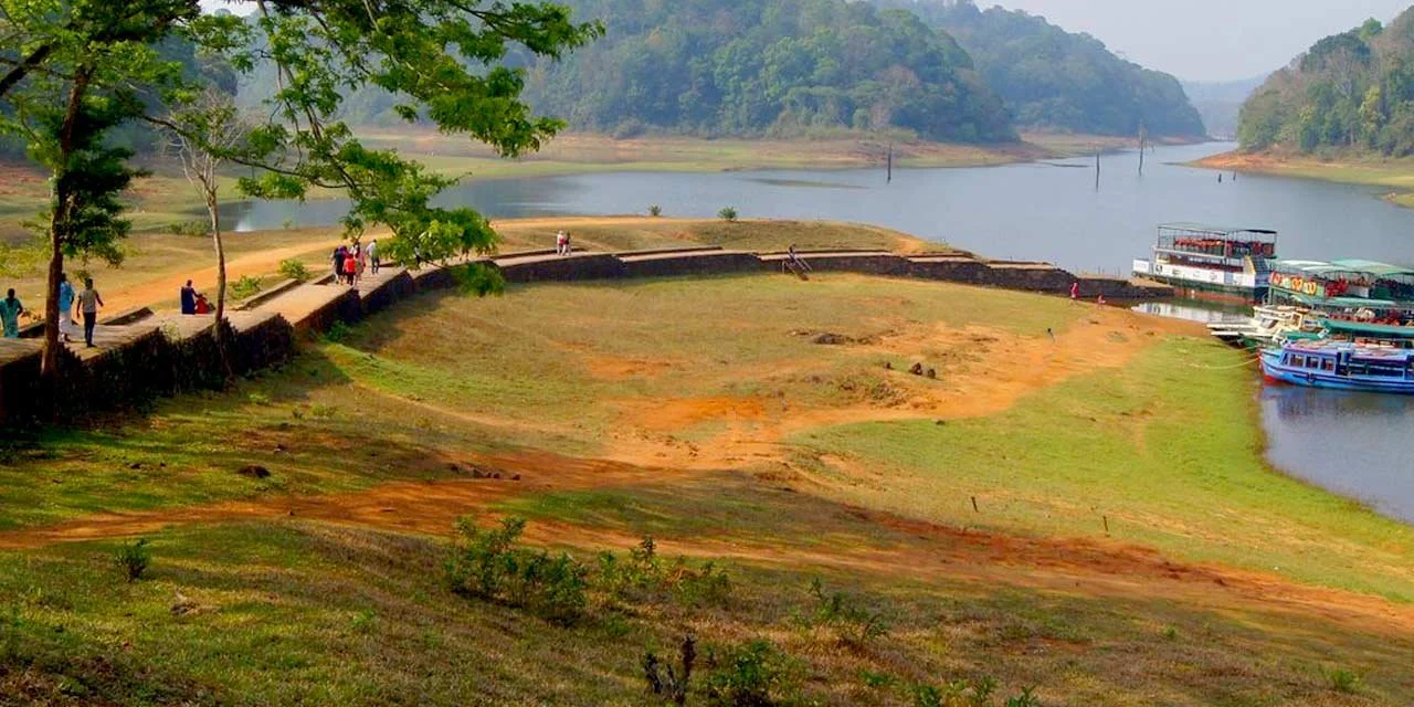View of a river with boats docked along the shore, surrounded by green hills and some trees, with a pathway along the riverbank where people are walking and standing.
