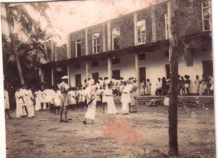 A black and white photo of children and adults outside a two-story building, with some children playing and others standing in line or gathering in groups.