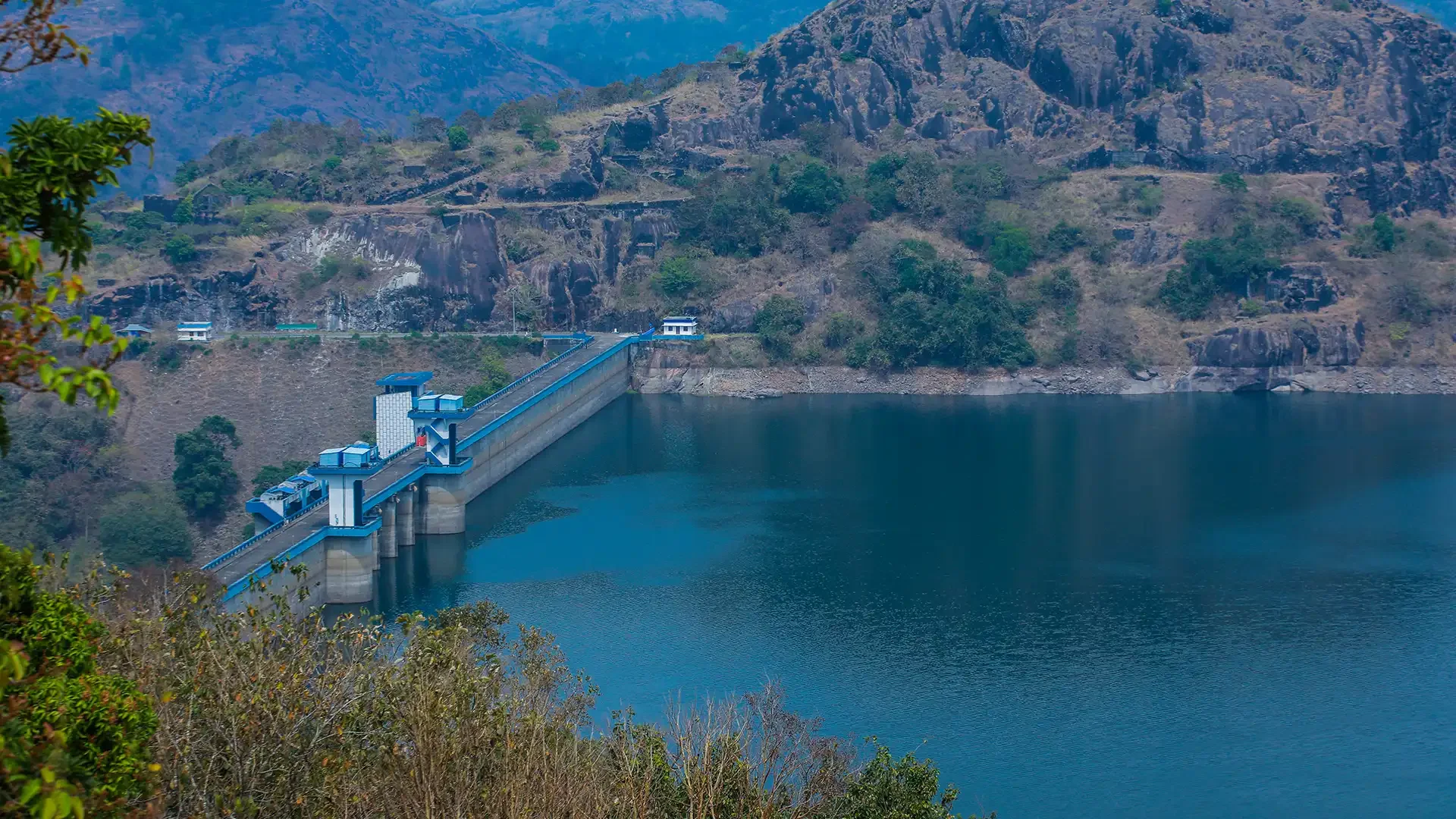 View of a dam with a blue spillway across a reservoir, surrounded by rocky hills and sparse vegetation.