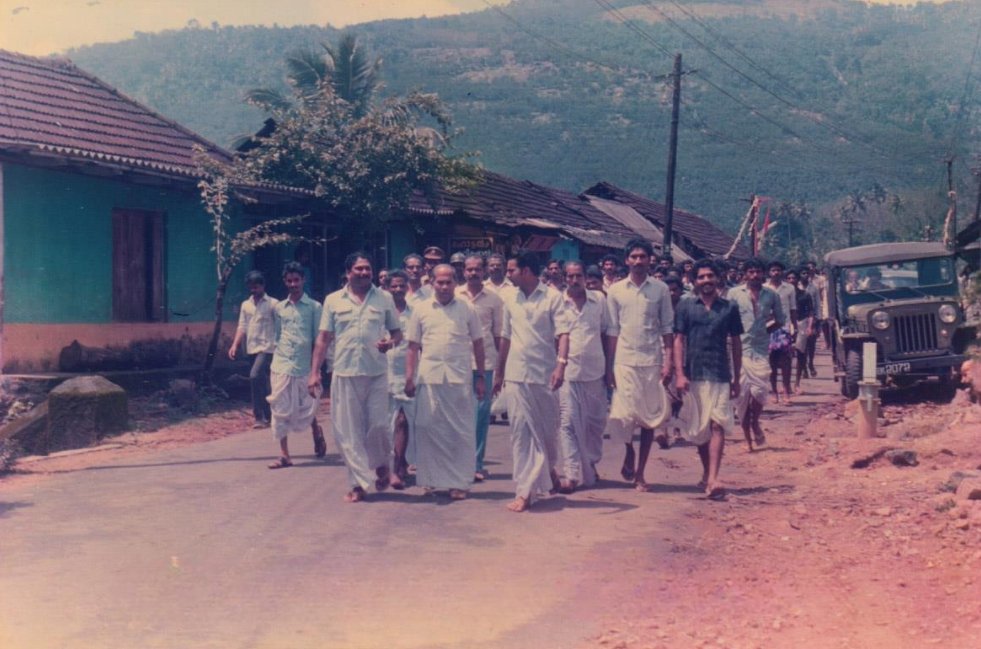 A group of men walking down a rural street, some wearing traditional white garments, with houses, a tree, and a vehicle in the background.