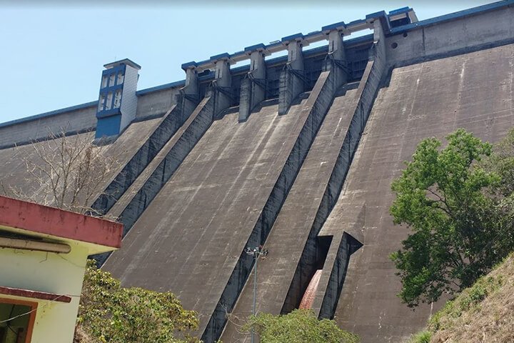 A large concrete dam with spillways and sluice gates, with trees and a building in the foreground.
