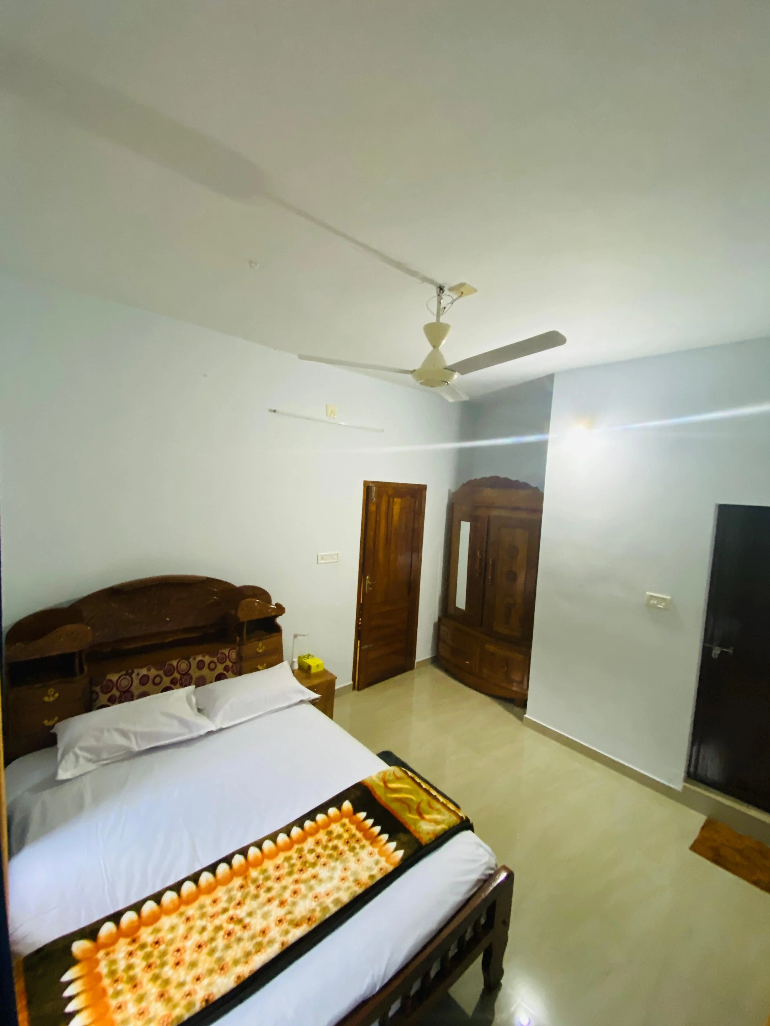 A simple bedroom with a wooden bed, white sheets, and a colorful blanket. There is a wooden wardrobe and a door, with light-colored tiled flooring and white walls. A ceiling fan hangs from the ceiling.