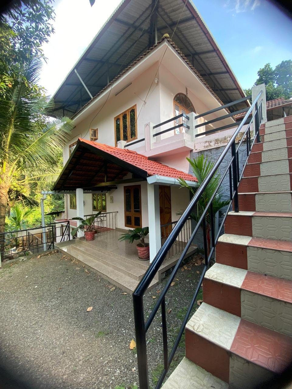 A two-story house with a front porch and stairs, surrounded by tropical plants, with a blue sky background.