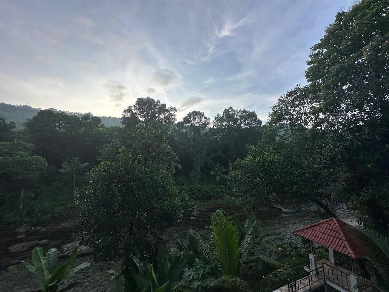 Lush green landscape with dense trees, a flowing river, and a small red-tiled roof structure in the foreground, under a partly cloudy sky.