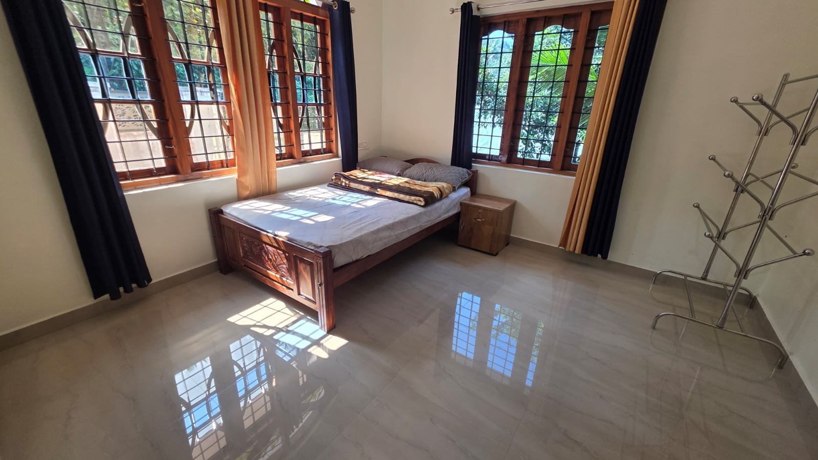 A bedroom with a wooden bed, two windows with blinds, a small wooden side table, a coat rack, and colorful curtains. Sunlight streams through the windows, reflecting on the tiled floor.
