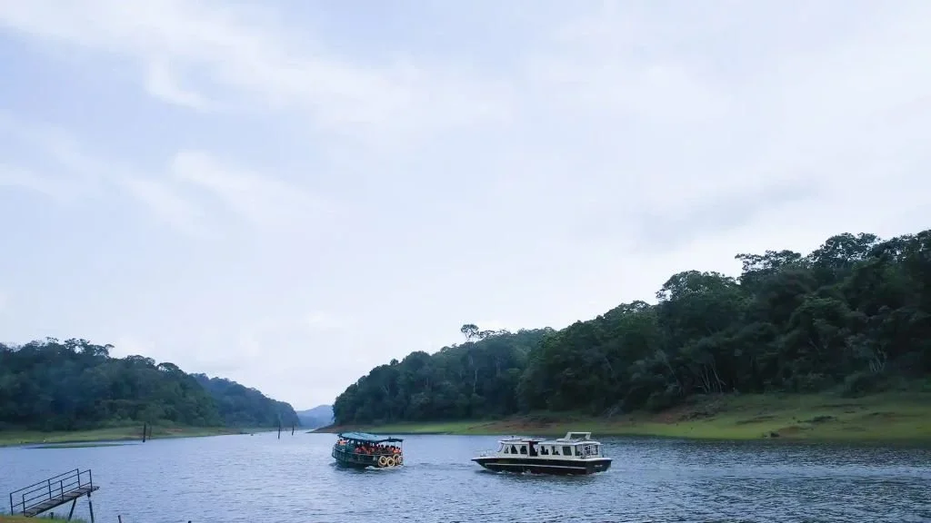 Two boats sailing on a wide river surrounded by lush green trees on both sides under a partly cloudy sky.