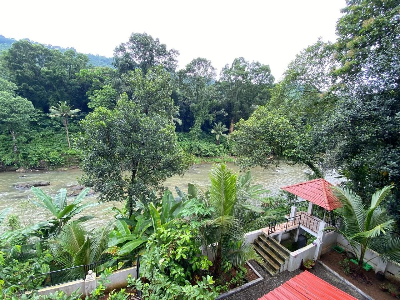 Lush green tropical landscape with a river, dense trees, and a small porch area with a red-tiled roof.