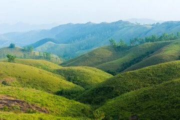 Lush green rolling hills with distant mountain ranges under a light blue sky.