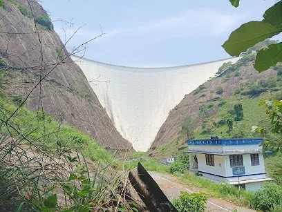 Dammed river valley with a large concrete dam, surrounded by green hills, and a small building with a blue roof in the foreground.
