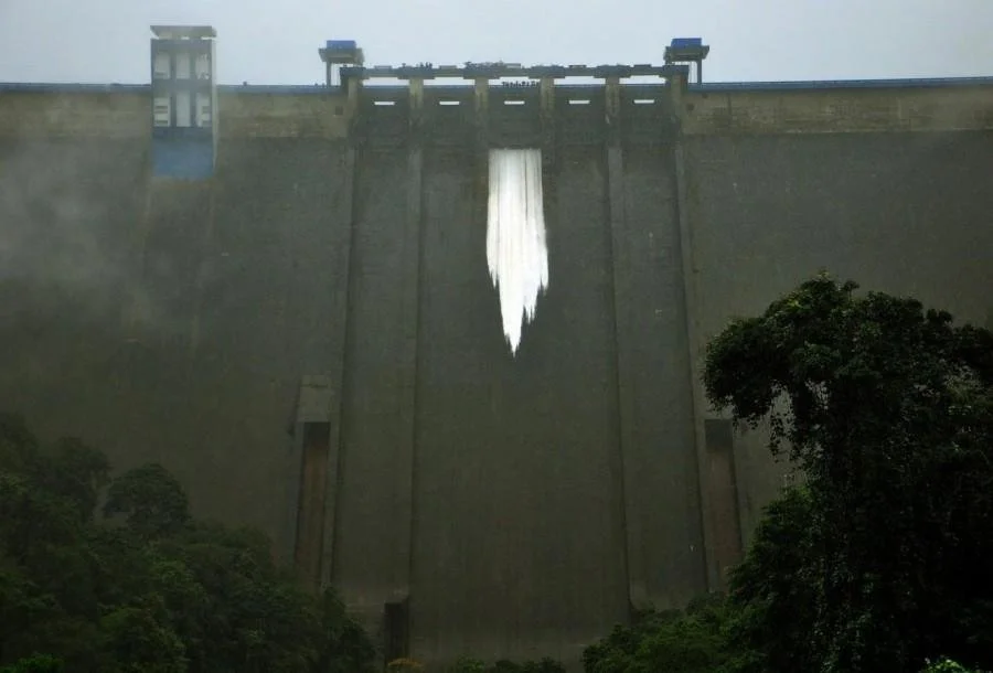 Large concrete dam with water spilling over the top, creating a white waterfall, surrounded by trees and a cloudy sky.