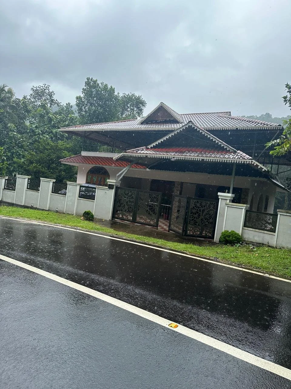 A house with a traditional roof design, surrounded by a white fence, on a rainy day with wet pavement and dark cloudy sky.