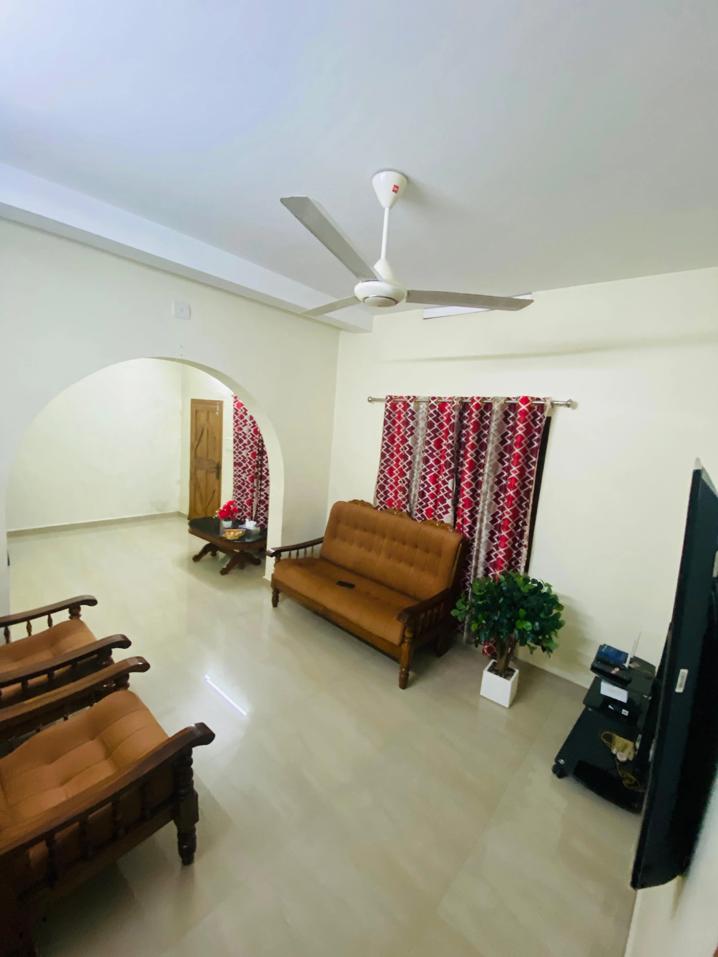 Living room with wooden sofa set, brown cushions, red and beige patterned curtains, a small table with flowers, a potted plant, a TV, and a ceiling fan.